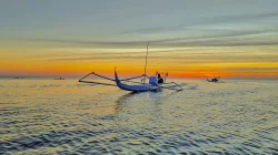 Perahu cadik nelayan melaju di laut tenang saat matahari terbenam dengan langit jingga