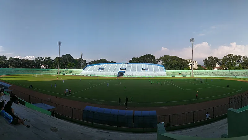 Arema FC Pulangkan Hanif Sjahbandi dari Persija Panorama Stadion Gajayana dengan tribun biru putih di seberang lapangan dan beberapa orang bermain bola di bawah langit biru