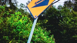 Rambu kuning tanjakan curam miring di tepi jalan yang dikelilingi semak dan pepohonan