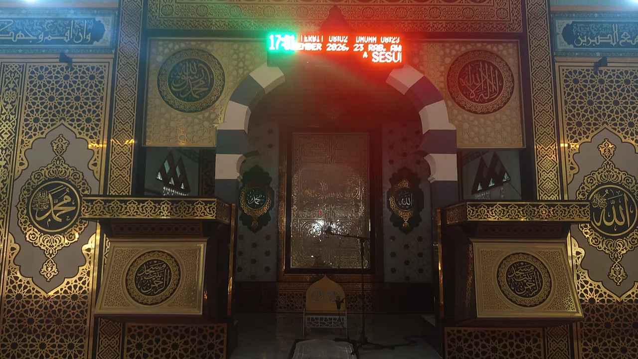 Interior masjid dengan mihrab berukir, kaligrafi emas, dan papan jadwal salat digital menyala