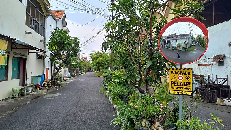 Bupati Gowa Tersangka Dugaan Pemerasan, Aset Rp5,5 M Disita Jalan lingkungan perumahan beraspal dengan deretan rumah, tanaman dalam pot, cermin cembung lalu lintas, dan papan peringatan persimpangan