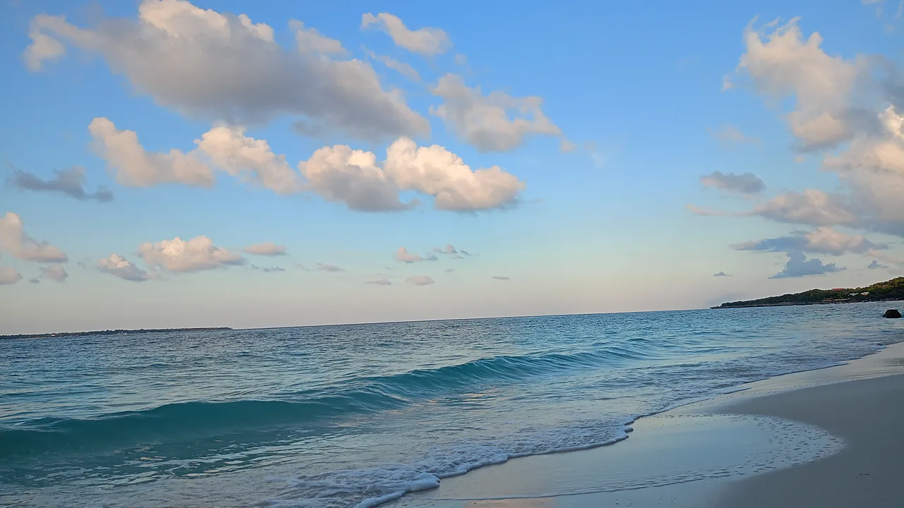 Ombak kecil memecah di pantai berpasir putih dengan laut biru toska dan langit biru berawan tipis
