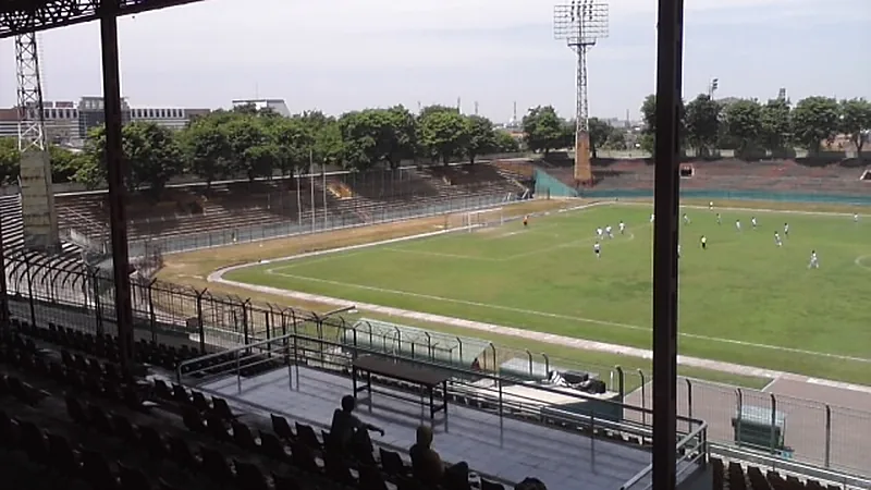 De Red FC vs RANS Nusantara, Aji Santoso Buru Poin Perdana Lapangan Stadion Gelora 10 November Tambaksari dilihat dari tribun beratap, dengan menara lampu dan pepohonan di belakang tribun seberang