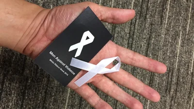 Telapak tangan terbuka memegang kartu hitam bertuliskan Men Against Violence dan pita putih simbol penolakan kekerasan terhadap perempuan