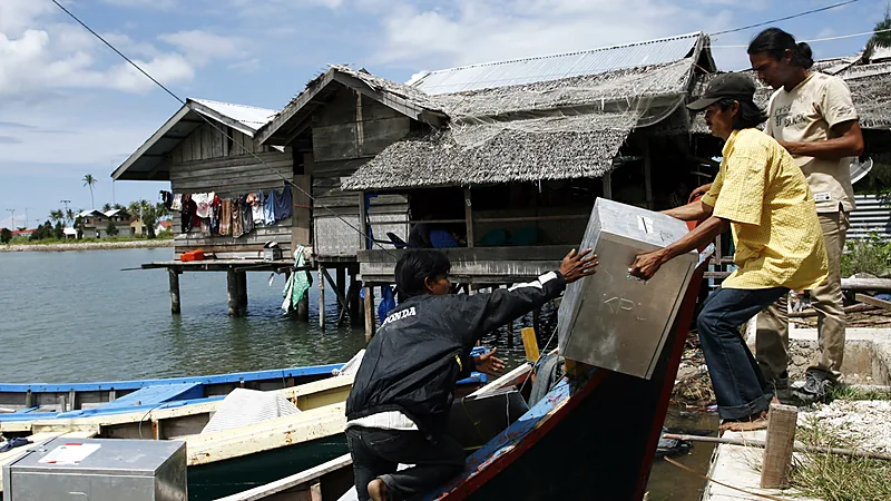 DPRD Bulukumba 2024-2029: 40 Kursi dan Pimpinannya Dua warga menurunkan kotak suara logam bertanda KPU dari perahu kayu di tepi air, dengan rumah panggung beratap seng dan nipah di latar belakang