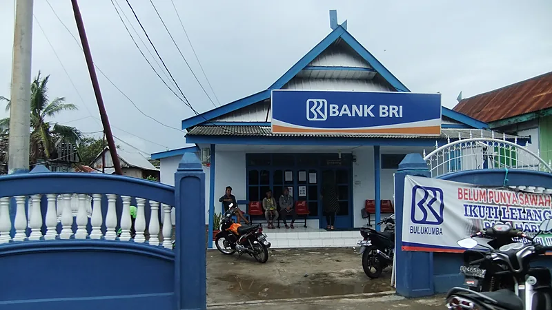 Kantor unit Bank BRI berupa bangunan bercat biru dan putih beratap pelana dengan papan nama besar, beberapa orang duduk di teras, dan sepeda motor terparkir di halaman