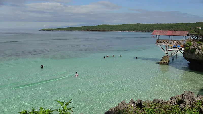 Perairan dangkal berwarna hijau toska di Tanjung Bira dengan beberapa orang berenang, tebing karang dan bangunan berdermaga kayu di sisi kanan