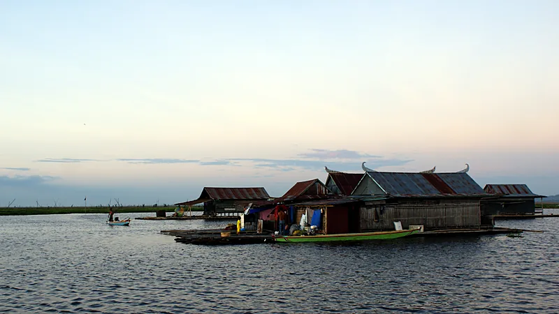 Festival Danau Tempe Digelar 23 September-5 Oktober Beberapa rumah rakit beratap seng dengan ujung atap melengkung mengapung di permukaan Danau Tempe menjelang senja, sebuah perahu hijau tertambat di sisinya dan perahu kecil lain melintas di kejauhan