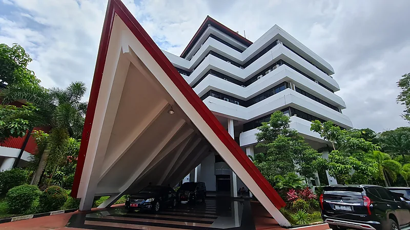 Wakil Rektor PTN se-Indonesia Bahas Kecurangan Seleksi dan AI di Unhas Gedung rektorat Universitas Hasanuddin bertingkat dengan kanopi segitiga merah putih di pintu masuk dan mobil terparkir di bawahnya