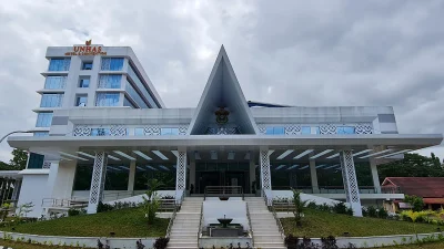 Gedung Unhas Hotel and Convention berwarna putih dengan atap segitiga tinggi di tengah, tangga lebar di depan, dan langit mendung