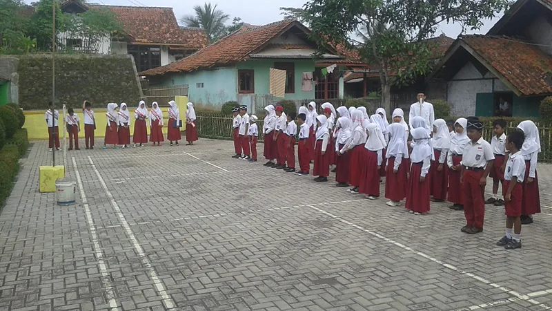 Siswa SDN 160 Tarampang Juara Mendongeng FTBI Bontobahari Murid sekolah dasar berseragam putih merah berbaris rapi membentuk huruf U di halaman paving sekolah dengan rumah beratap genteng di latar belakang