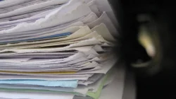 Tumpukan tebal lembaran kertas dan berkas dokumen berbagai warna yang dijepit dalam ordner, difoto dari samping
