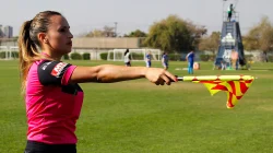 Asisten wasit perempuan berkaus merah muda merentangkan bendera bermotif kuning merah ke samping di tepi lapangan sepak bola