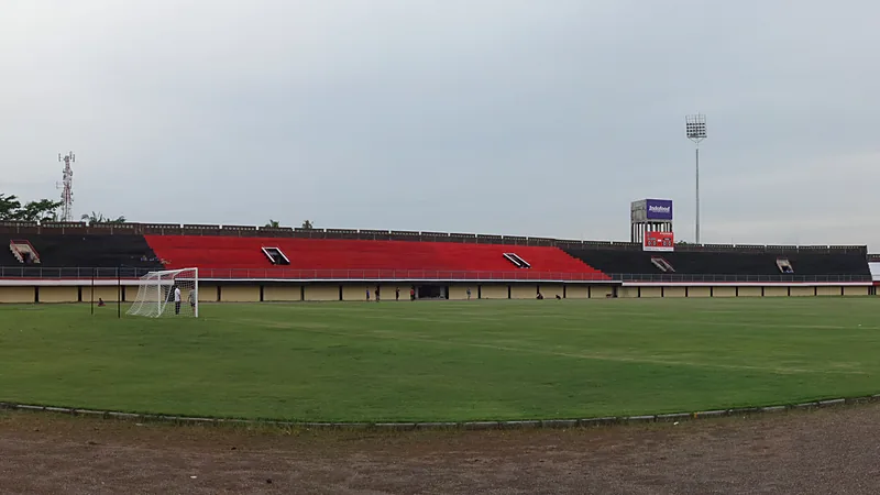 Tribun bercat merah dan hitam Stadion Kapten I Wayan Dipta yang kosong dengan lapangan hijau dan gawang di sisi kiri