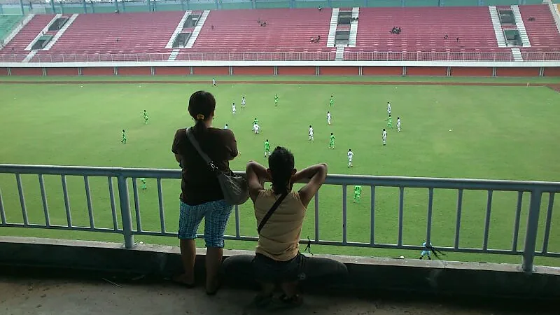 Dewa United Tekuk PSS Sleman 2-1, Kudeta Puncak Klasemen Dua penonton berdiri di balik pagar tribun menyaksikan pertandingan sepak bola di lapangan Stadion Maguwoharjo yang berkursi merah