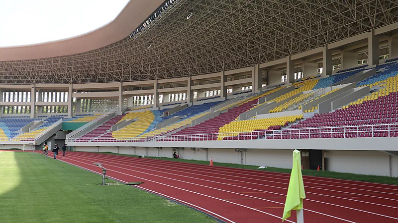 Tribun Stadion Manahan dengan kursi merah, kuning, dan biru di bawah atap rangka baja, dilihat dari tepi lintasan atletik merah