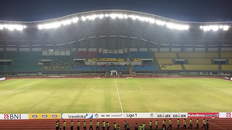 Lapangan Stadion Patriot Chandrabhaga pada malam hari dengan lampu sorot menyala dan tribun berkursi kuning, biru, dan merah yang sebagian terisi