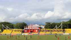 Tribun timur Stadion Haji Agus Salim berkursi kuning dengan atap runcing khas Minangkabau di tengahnya, pertandingan sepak bola berlangsung di lapangan