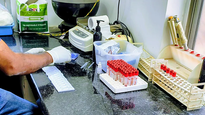 Tangan bersarung lateks putih menyiapkan alat tes cepat di meja laboratorium, dengan rak tabung sampel bertutup merah, pipet, dan kotak jarum suntik sekali pakai