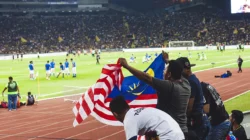 Suporter membentangkan bendera di tribune stadion saat pemain berlatih di lapangan