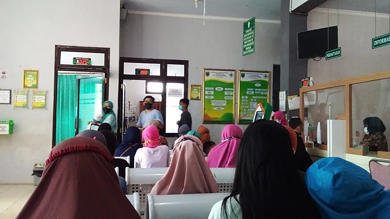 Ruang tunggu puskesmas yang penuh, pasien duduk berderet di kursi besi menghadap loket, dengan papan penunjuk pendaftaran dan informasi terpasang di dinding