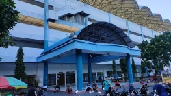 Pintu masuk utama Stadion Maguwoharjo berkanopi biru dengan atap rangka baja melengkung di atasnya dan pengunjung duduk di pelataran