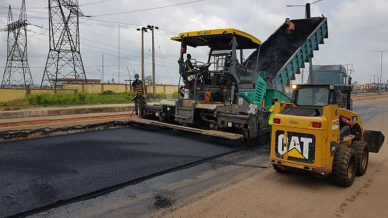 Jalan Tanete-Tanaberu Bulukumba Mulai Mulus dan Melebar Mesin penghampar aspal kuning dan truk pengangkut sedang menghamparkan lapisan aspal hitam baru di badan jalan, didampingi sebuah loader kecil