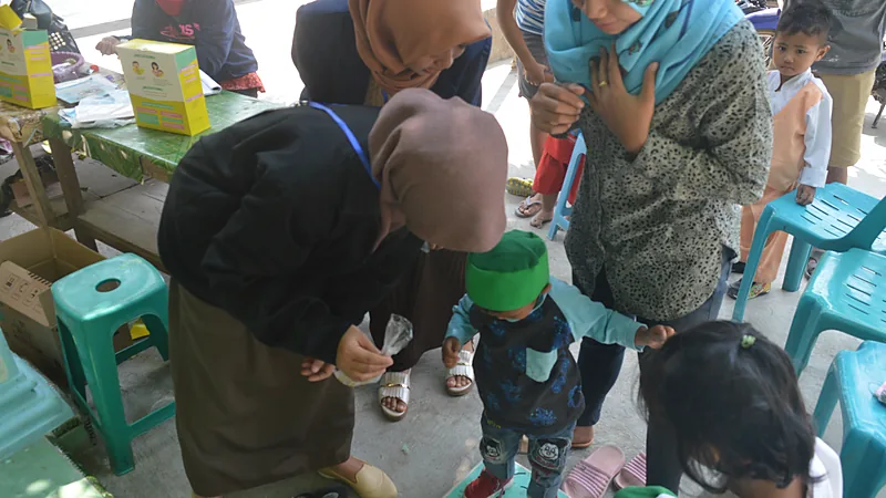 Kader posyandu melayani balita dan ibu yang mengantre di bawah atap terbuka dengan meja layanan, kursi plastik dan kotak obat di belakangnya