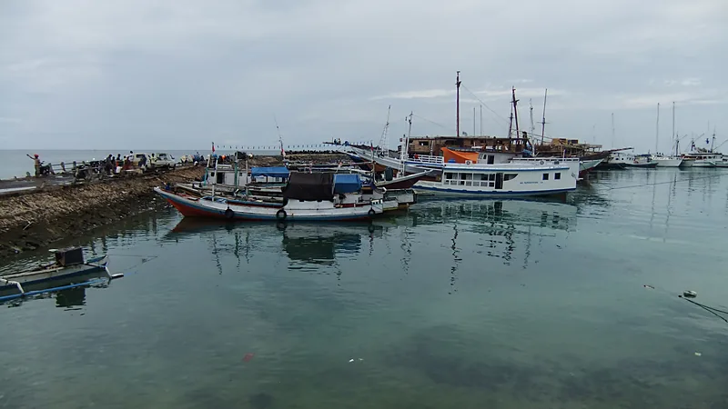 Profil Kecamatan Bonto Bahari: Bira, Ara, Tanah Beru Perahu-perahu nelayan dan kapal kayu bertambat di perairan dangkal Pelabuhan Bira, dengan dermaga batu memanjang di sisi kiri dan langit mendung di atasnya