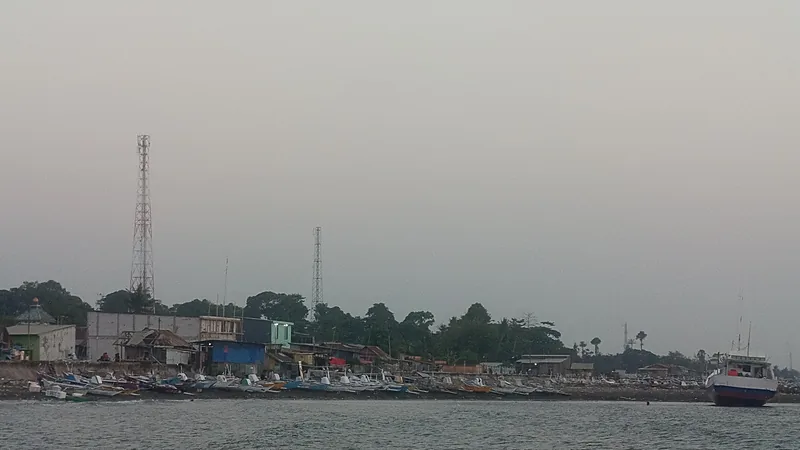 Profil Kecamatan Bontotiro Bulukumba: 12 Desa, 1 Kelurahan Deretan perahu nelayan berkatir bersandar di tepi pantai berbatu di depan rumah dan bangunan rendah, dua menara telekomunikasi menjulang di antara pepohonan, dengan laut kelabu dan langit berkabut di latar