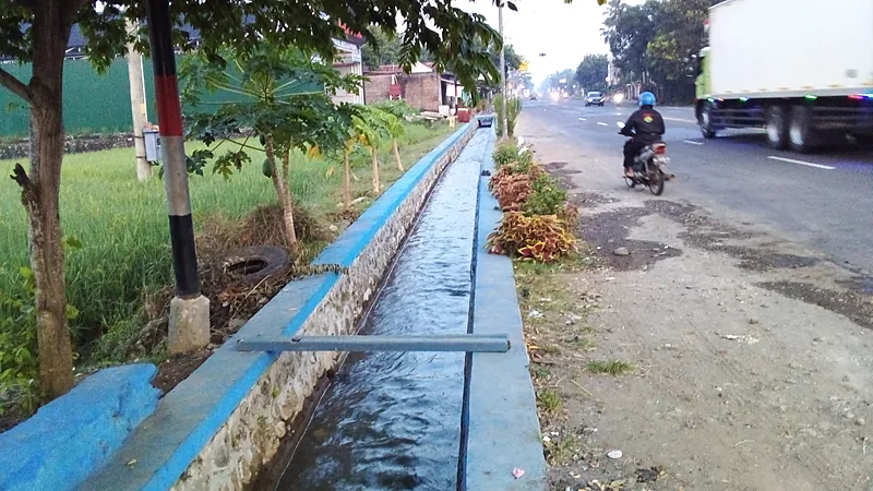 Kemenham Sulsel Petakan Keluhan Bansos Warga Bontomanai Saluran irigasi beton bercat biru mengalirkan air di antara sawah dan jalan raya, seorang pengendara motor melintas dari belakang