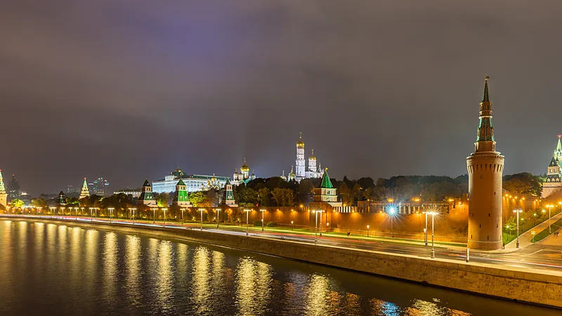 Pemandangan malam tembok dan menara bata merah Kremlin Moskow yang diterangi lampu di tepi Sungai Moskwa, dengan kubah emas katedral dan istana putih di balik tembok serta pantulan cahaya di permukaan sungai