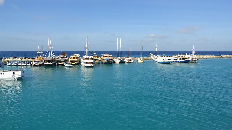 KMP Awu Awu Gantikan Balibo di Lintasan Bira–Pamatata Deretan kapal kayu bertiang dan kapal wisata putih berlabuh di kolam pelabuhan berair biru kehijauan yang dibatasi pemecah gelombang batu, dengan laut lepas dan langit cerah di belakang