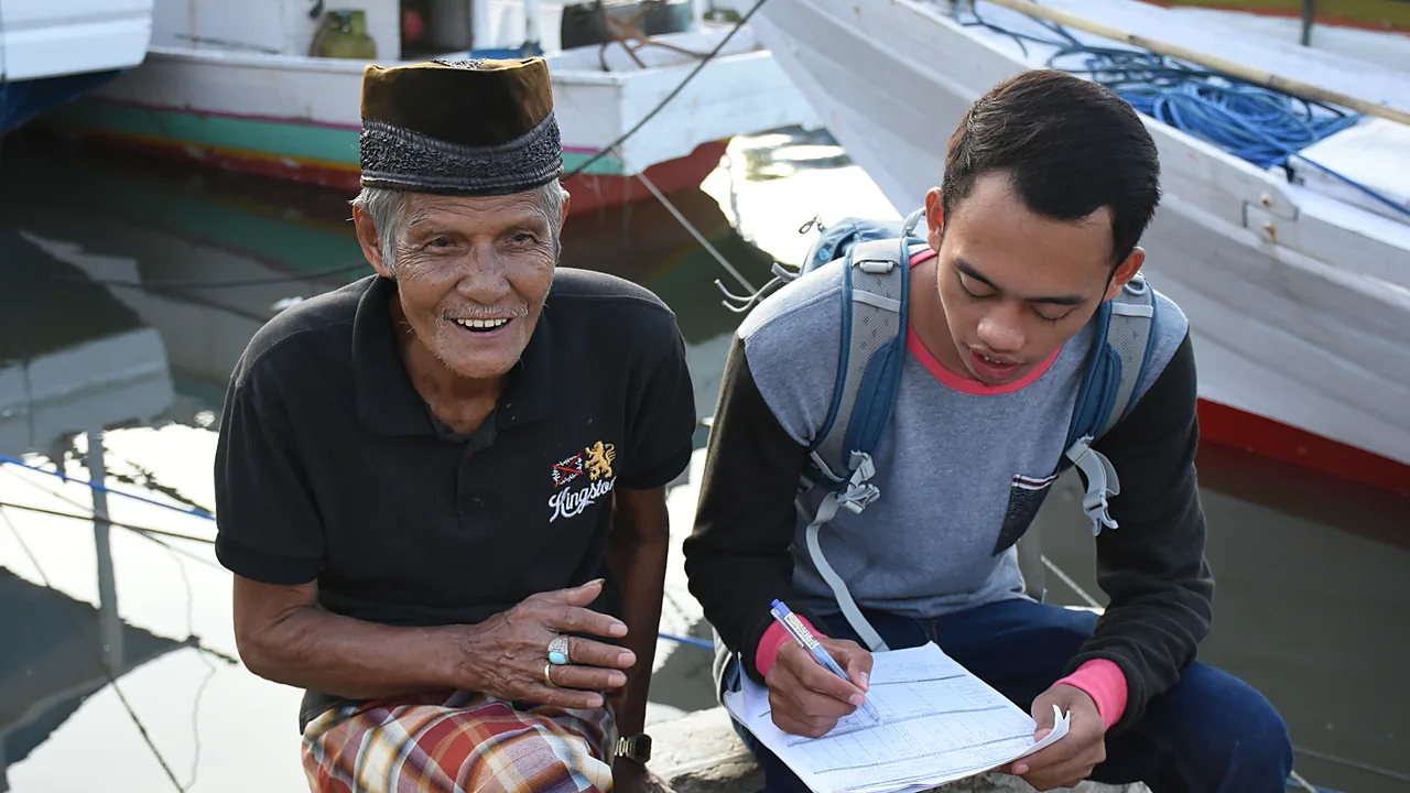 Seorang nelayan lanjut usia bersongkok dan bersarung duduk di tepi dermaga sambil tersenyum, didampingi petugas muda bertas punggung yang mengisi lembar pendataan, berlatar perahu kayu putih
