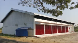 Bangunan gudang berdinding seng abu-abu dengan deretan rolling door merah dan terpal biru di sampingnya