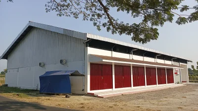 Bangunan gudang berdinding seng abu-abu dengan deretan rolling door merah dan terpal biru di sampingnya
