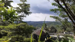 Pemandangan lembah hijau berbukit dengan atap gazebo di antara pepohonan rimbun dan langit berawan