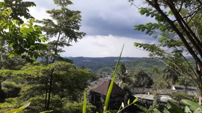Pemandangan lembah hijau berbukit dengan atap gazebo di antara pepohonan rimbun dan langit berawan