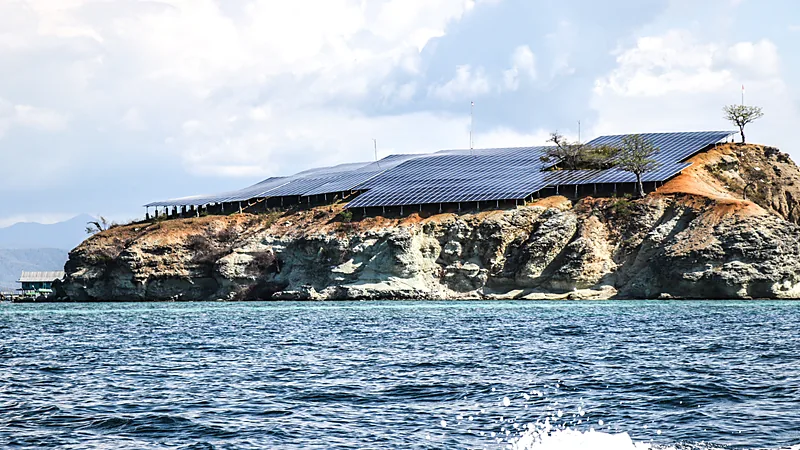 Deretan panel surya berwarna biru gelap menutupi puncak pulau karang kecil bertebing kapur di tengah laut biru, dengan langit berawan dan pegunungan samar di latar belakang