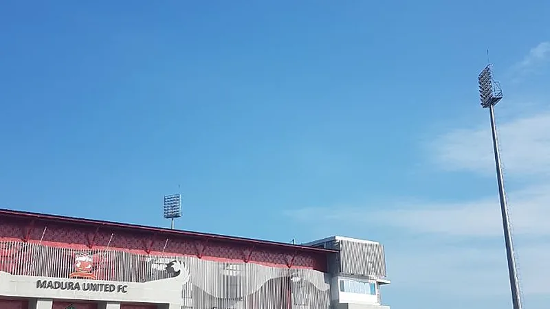 Bagian atas fasad Stadion Gelora Madura Ratu Pamelingan bertuliskan Madura United FC dengan menara lampu di bawah langit biru