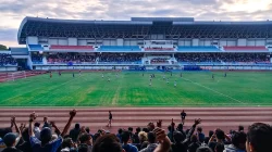 Pertandingan di Stadion Mandala Krida Yogyakarta dengan tribun beratap lengkung, dilihat dari belakang penonton yang mengangkat tangan