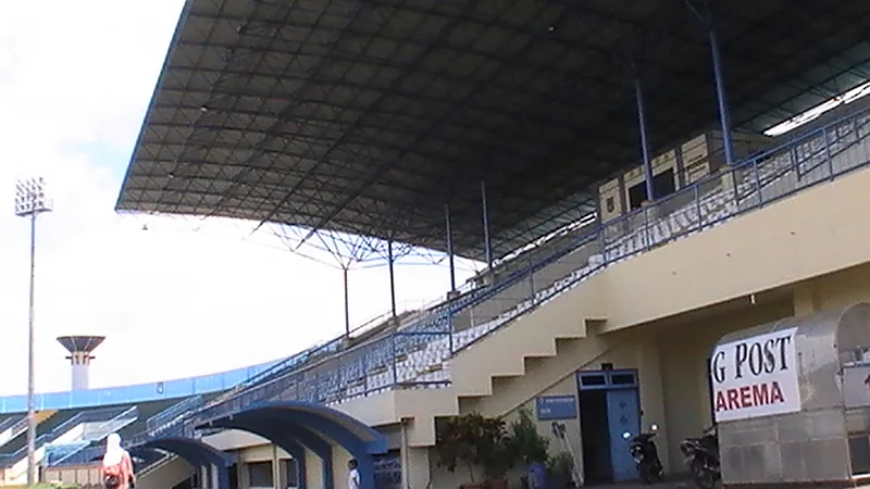 Tribun beratap baja Stadion Kanjuruhan dalam keadaan kosong pada siang hari, dengan papan bertuliskan Arema di dinding luar tribun