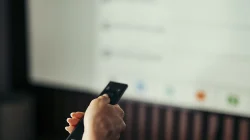 Tangan memegang remote menghadap layar televisi yang menampilkan menu aplikasi streaming