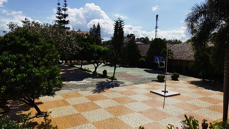 Halaman sebuah SMP negeri berpaving blok kuning putih dengan tiang bendera di tengah dan lapangan basket berlantai semen di belakangnya
