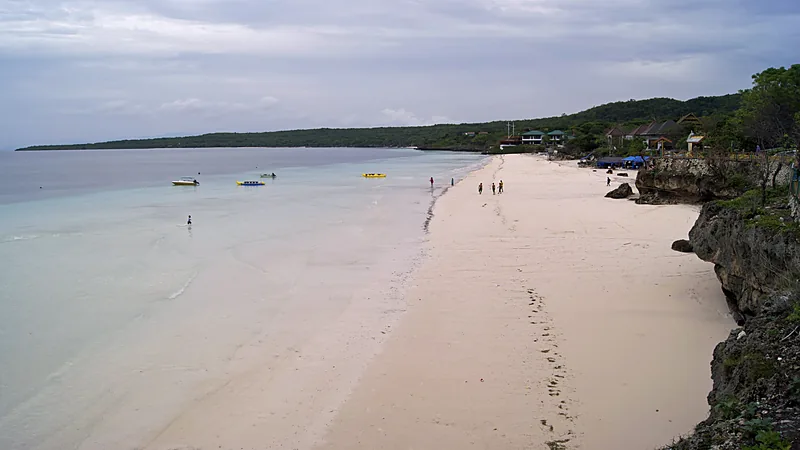 Hamparan pasir putih Pantai Tanjung Bira dengan beberapa perahu kecil di perairan dangkal, tebing karang rendah di sisi kanan, dan bangunan penginapan di kejauhan