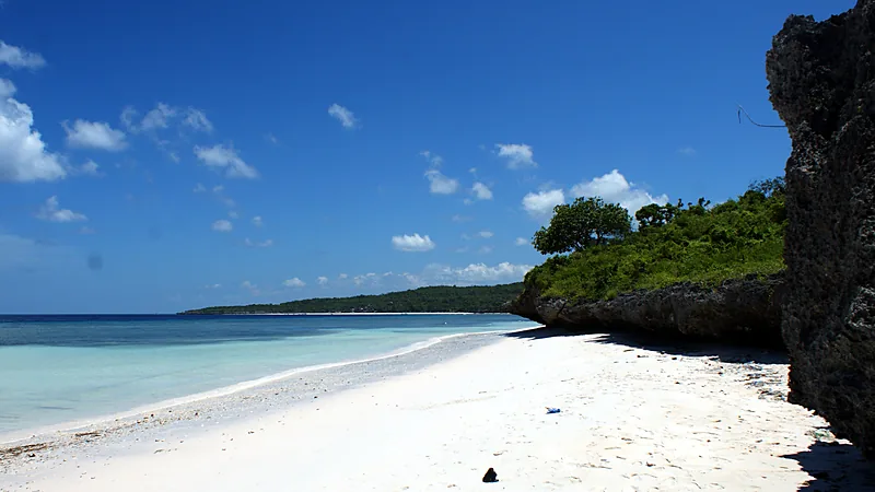 Tiket Masuk Pantai Bira Bulukumba: Tarif Perda dan Akses Pantai berpasir putih dengan air laut toska jernih, tebing karang hitam berpepohonan di sisi kanan, dan tanjung hijau di kejauhan di bawah langit biru