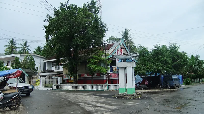 Terduga Pelaku Pelecehan Nakes di Gantarang Ditangkap Pertigaan jalan beraspal basah di Bulukumba dengan tugu putih bermahkota miniatur perahu, pohon rindang, dan sepeda motor terparkir