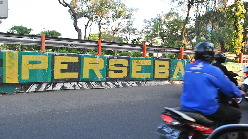 Mural bertuliskan Persebaya berwarna kuning di atas latar hijau pada dinding tepi jalan, dengan pengendara sepeda motor melintas