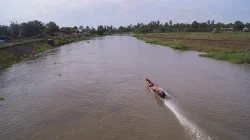 Perahu kayu bermotor melaju meninggalkan jejak buih di sungai cokelat yang lebar, diapit tepian berumput, sawah, pohon kelapa, dan rumah-rumah di bawah langit mendung