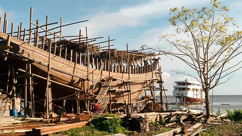 Lambung kapal kayu setengah jadi dikelilingi perancah dan tumpukan kayu di pesisir, dengan kapal putih berlabuh di laut dan pohon di sisi kanan